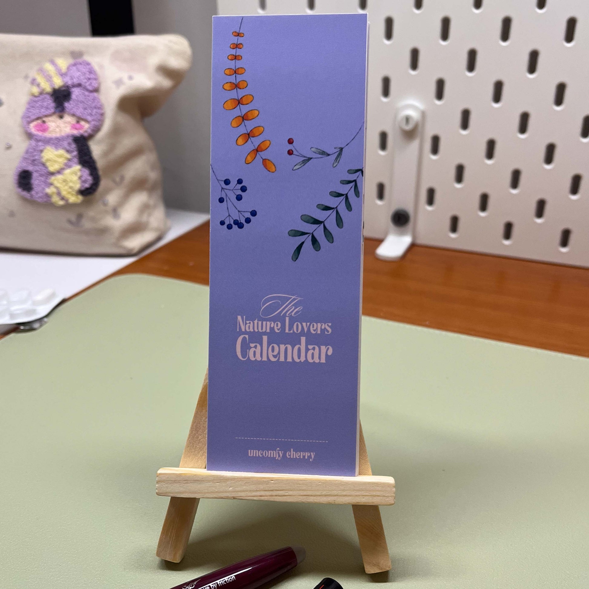Cute Uncomfy Cherry bookmark calendar displayed on a small wooden easel with illustrated monthly pages.