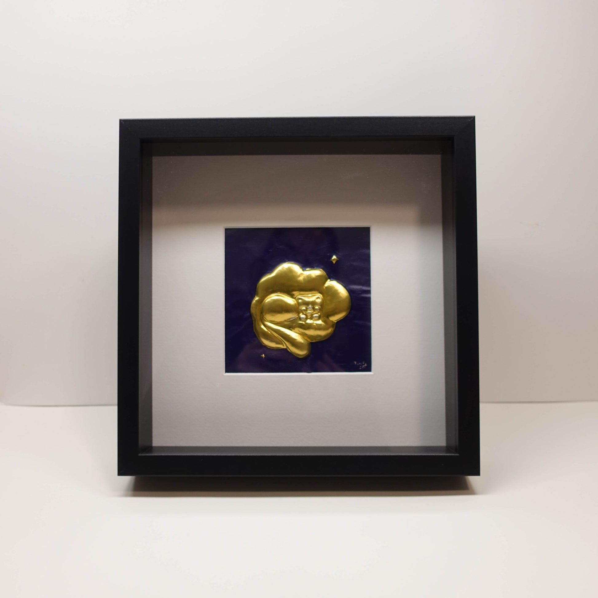 Brass 3D sculpture of a sleeping cat resting on clouds, set against a painted blue background in a recessed frame, 25 × 25 cm.