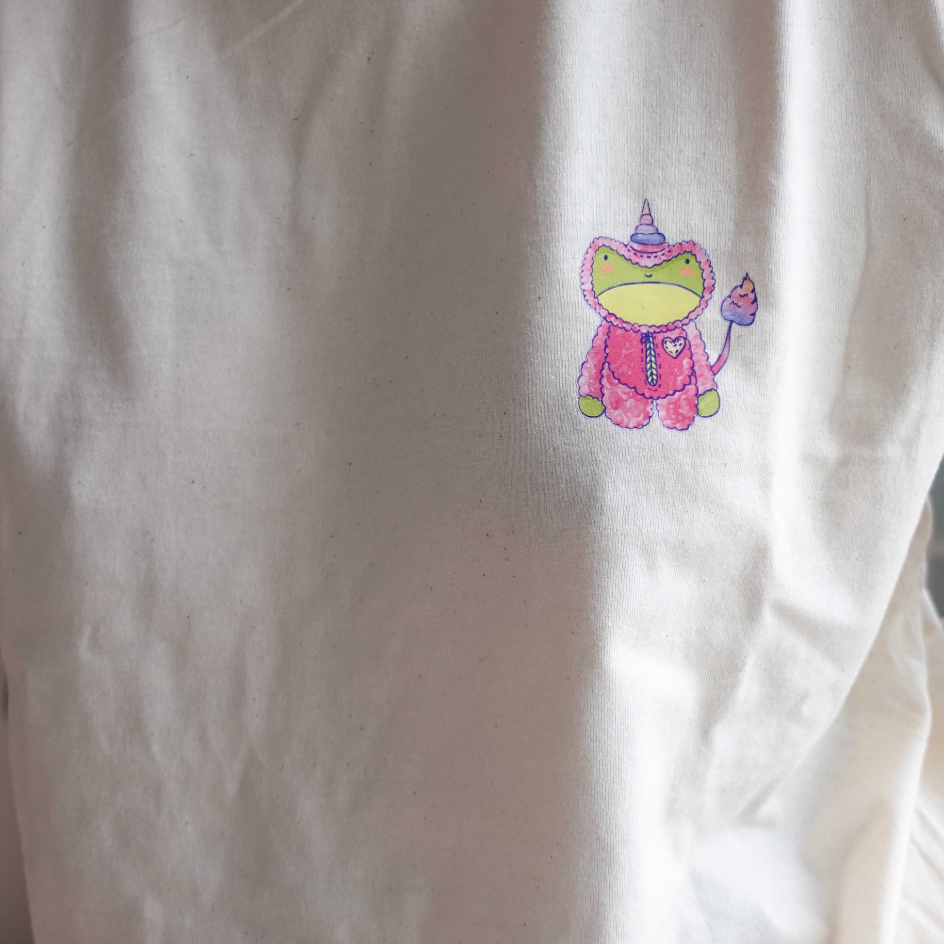 Front view and close up of the Fluff Club  t-shirt in natural raw color, featuring a original design by uncomfy cherry from the frog in a unicorn suit